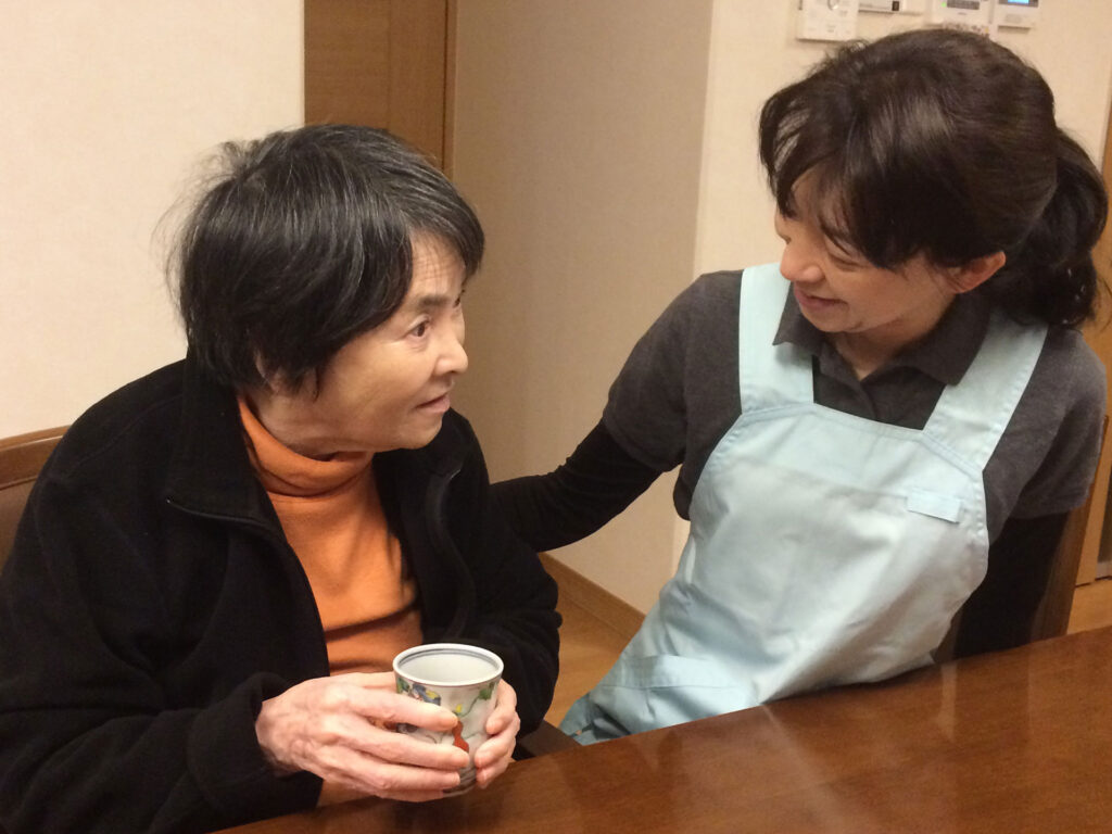 お茶を飲む高齢女性に寄り添い背中をさするスタッフ